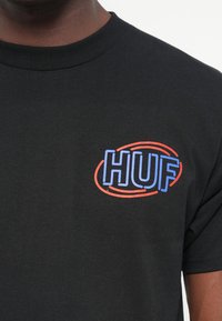Sort bomulds T-shirt med et farverigt "HUF" logo i blå og rød. Ribbet rund hals og korte ærmer. Glat tekstur.