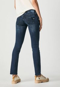 Mujer vestida con jeans ajustados azul oscuro y zapatillas beige con detalles de estampado animal, de pie frente a un fondo claro y liso.