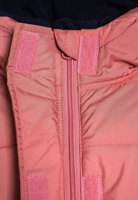 Doudoune rose avec fermeture éclair, patchs en velcro et texture matelassée. Doublure intérieure en polaire bleu marine. Extérieur lisse et résistant à l'eau.