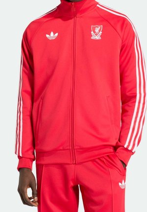 Osoba ubrana w czerwony dres Adidas z białymi paskami na rękawach i spodniach, herb Liverpool Football Club na piersi kurtki.