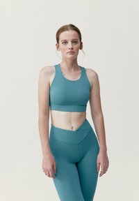 Teal urheiluliivit ja korkeavyötäröiset legginsit sileästä, venyvästä kankaasta. Liivissä on leveä vyö, ja legginsseissä on taitettava vyötäröyksityiskohta.