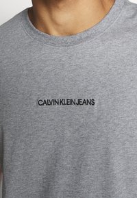 Camiseta de algodón gris con cuello redondo y mangas cortas, con el texto bordado en negro "CALVIN KLEIN JEANS" en la parte frontal.