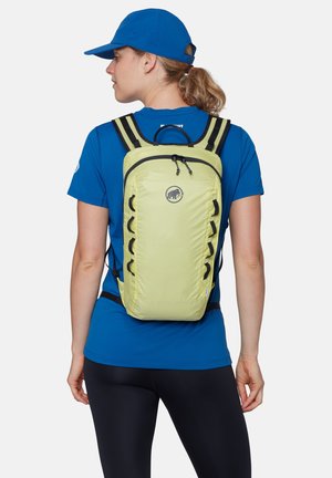 Femme portant une casquette et une chemise bleues, avec un sac à dos compact vert clair à bretelles noires, debout sur un fond blanc uni.