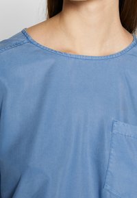 Lys blå bomuldsbluse med rund halsudskæring, synlige sømme og en brystlomme. Stoffet virker glat og let tekstureret.