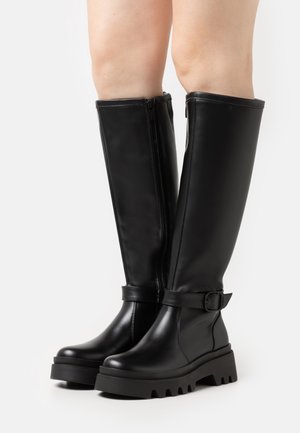 Sorte kneehøye støvler laget av glatt syntetisk lær, med glidelås på siden, en bred stropp med spenne, og kraftige gummisåler.