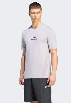 Ung mand iført en lysegrå kortærmet sportstrøje med AMG-logo og mørkegrå cargoshorts, der står mod en ensfarvet baggrund.