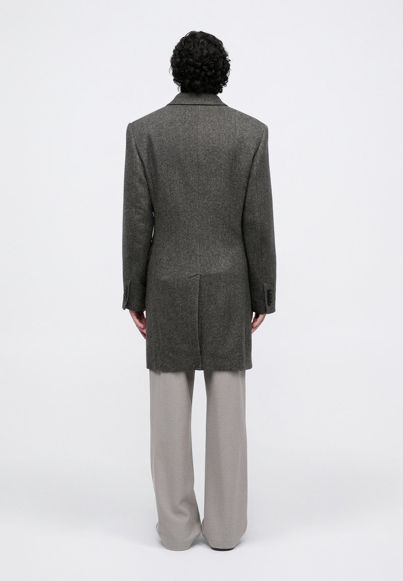 Un homme avec des cheveux bouclés se tient de dos, portant un long manteau gris et un pantalon large gris clair, sur un fond blanc uni.