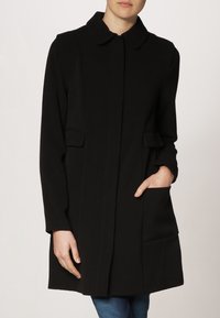 Černý kabát vyrobený z měkké látky, s límcem, dvěma předními kapsami a rovnou siluetou. Hladký povrch, minimální šití.