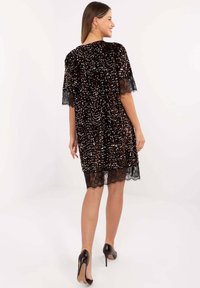 Czarna sukienka z cekinami, z przezroczystymi koronkowymi rękawami i dołem, o długości do kolan i luźnym fasonie, uzupełniona brązowymi butami na wysokim obcasie.