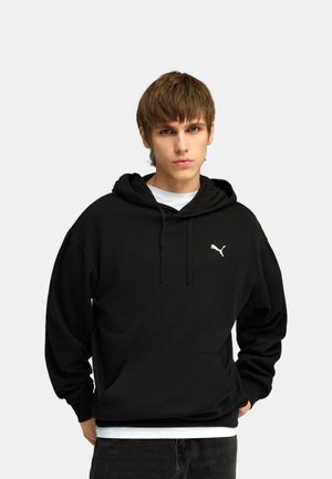 Mladý muž v černé mikině Puma s kapucí, šňůrkami a klokaní kapsou, stojící před jednoduchým světlým pozadím.