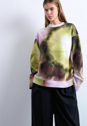 Jeune femme portant un sweat-shirt ample tie-dye aux tons verts, marron et roses, avec un pantalon noir à jambes larges, sur un fond blanc uni.