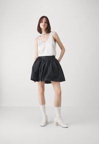 Top in satin bianco con scollatura profonda a V abbinato a una gonna nera a palloncino. La modella indossa stivali bianchi con tacco a blocco. Sfondo minimalista.