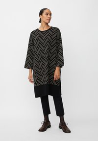 Černá tunika se zlatým vzorem chevron, dlouhé rukávy a boční rozparky, spárovaná s černými kalhotami a texturovanými hnědými botami.