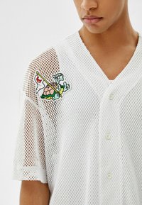 Valkoinen verkkomainen lyhythihainen paita, jossa on V-aukko ja napitus. Vasemmassa olkapäässä on värikäs kilpikonnakuvioinen kangasmerkki.