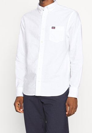 Hombre con camisa blanca abotonada con un bolsillo en el pecho que muestra un pequeño logo de GANT, combinado con pantalones azul marino oscuro.