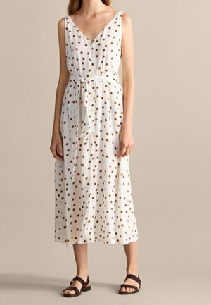Robe midi blanche sans manches à pois marron, col en V, boutonnée à l'avant, ceinture nouée à la taille, portée avec des sandales noires sur fond beige.