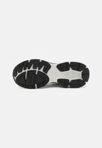 Sorte såle til sneakers i svart-hvitt med dype spor, teksturert gummi, og et emblem. Har et hevet sentralt design for grep og støtte.