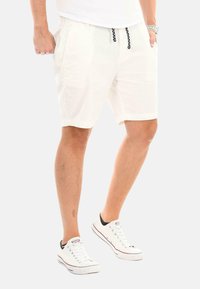 Weiße Baumwollshorts mit elastischem Bund, schwarzem Kordelzug und Seitentaschen. Kombiniert mit weißen Sneakers mit schwarzen Akzenten.