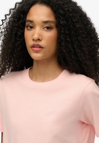 Różowa bawełniana koszulka z okrągłym dekoltem i krótkimi rękawami. Posiada subtelną haftowaną aplikację "SUPERDRY" na piersi. Gładka faktura, luźny krój.