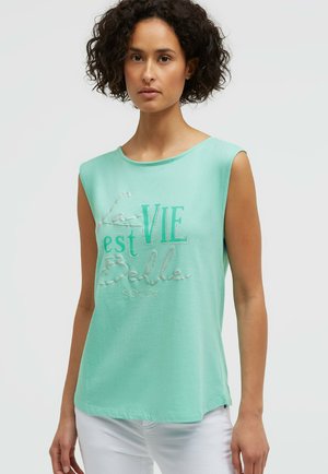 Frau mit lockigem Haar, die ein ärmelloses mintgrünes Oberteil mit dem Schriftzug „La Vie est Belle“ und eine weiße Hose trägt, vor einem schlichten hellen Hintergrund.