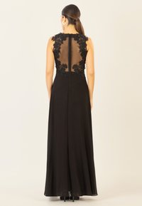 Langes schwarzes Kleid mit einem transparenten Rücken und floralem Spitzenappliken, ärmellosem Design und fließendem Stoff, der bis zum Boden reicht.