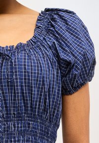 Μπλε σκούρο μπλούζα με λευκό σχέδιο πλέγματος, λεπτομέρεια ρυτίδωσης στη λαιμόκοψη, κοντά φουσκωτά μανίκια και συγκεντρωμένη λεπτομέρεια στη μέση σε μαυρισμένο δέρμα.