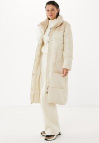 Lång krämfärgad pufferjacka med en mjuk beige krage, dragkedja fram och sidofickor. Den har en kviltad textur och detaljer i guldton.