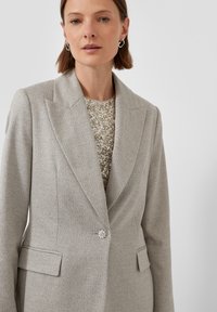 Vrouw in een lichtgrijs op maat gemaakt blazer met een glinsterende zilveren top eronder, die zilveren creolen draagt, tegen een effen witte achtergrond.
