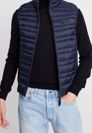 Homme portant un gilet matelassé bleu marine sur une chemise noire à manches longues et un jean bleu clair, une main dans la poche.