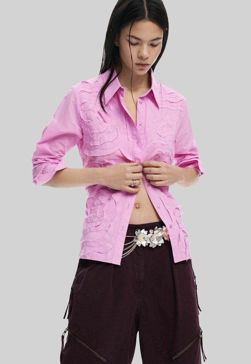 Roosa nööbitav särk, millel on tekstuuriga lilleline tikand, pikkade varrukatega, mille varrukad on üles keeratud, seljas tume burboonivärvi pükste peal, millel on dekoratiivsed vöödetailid.