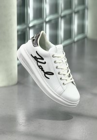 Shoe branca de Karl Lagerfeld com script preto "Karl" na lateral e detalhe preto no calcanhar, suspensa acima de um chão cinza perto de blocos de vidro.