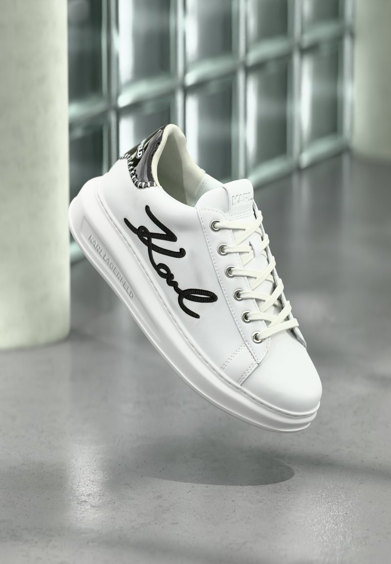 Shoe branca de Karl Lagerfeld com script preto "Karl" na lateral e detalhe preto no calcanhar, suspensa acima de um chão cinza perto de blocos de vidro.
