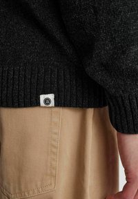 Schwarzer Strickpullover mit geripptem Saum, Etikett sichtbar. Kombiniert mit khakifarbenen Hosen, die einen strukturierten Stoff und Details an den Seitentaschen zeigen.