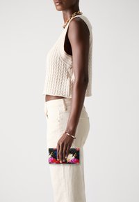 Kukka-aiheinen musta lompakko kädessä, jossa on eloisia värejä. Käyttäjällä on päällään vaalea neulottu crop-top ja vaaleat housut.