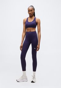 Αθλητική γυναίκα με μοβ αθλητικό σουτιέν και κολάν με λευκά λογότυπα Puma, λευκές κάλτσες και λευκά αθλητικά παπούτσια, που στέκεται μπροστά σε απλό φόντο.