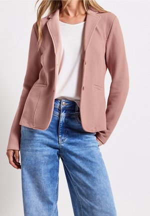 Frau trägt einen strukturierten rosa Blazer, ein weißes T-Shirt und hoch taillierte blaue Jeans, posiert mit locker hängenden Händen an den Seiten.