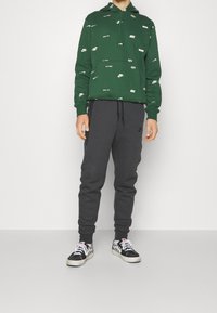 Vihreä huppari, jossa on hajallaan valkoisia logoja, yhdistettynä tummanharmaisiin collegehousuihin ja mustiin lenkkareihin, joissa on valkoinen ruutukuvio.