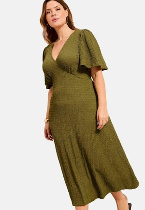 Frau trägt olivgrünes, strukturiertes Midikleid mit Puffärmeln und V-Ausschnitt, steht vor einem schlichten hellen Hintergrund.