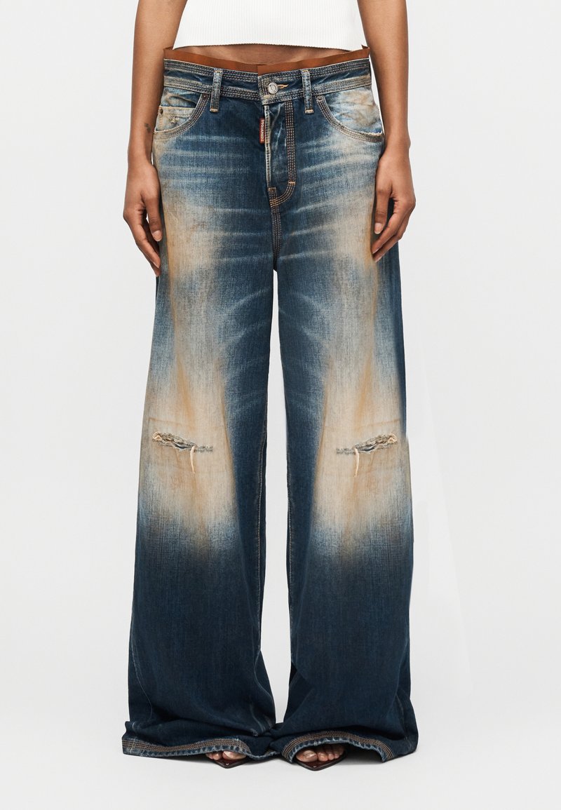 Weit geschnittene, ausgewaschene blaue Jeans mit ausgefransten Knien, getragen von einer Person mit freiem Oberkörper und Sandalen, die vor einem einfarbigen Hintergrund steht.