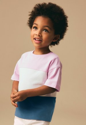 Lachend jong kind met krullend haar, gekleed in een pastelroze, wit en blauw shirt, kijkt iets omhoog tegen een beige achtergrond.