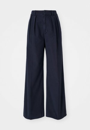 Un pantalon large bleu marine en coton, avec des plis à l'avant, une taille haute et une fermeture éclair avec un bouton.