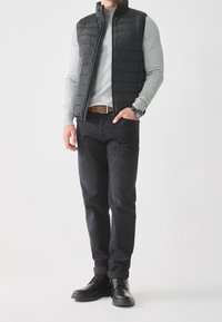 Black padded vest over a light gray sweater, dark jeans, and black shoes. Features a smooth texture and a fitted design. Brown belt included.