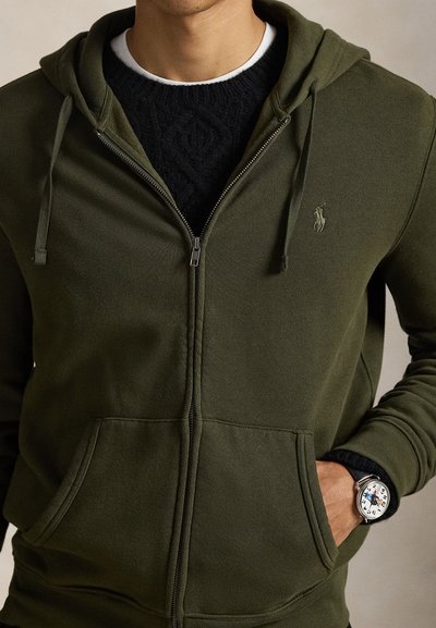 Φούτερ με κουκούλα και φερμο�υάρ σε ελιά χρώμα, με κορδόνια, εμπρόσθιες τσέπες και μικρό λογότυπο. Φορεμένο πάνω από ένα σκούρο πλεκτό πουλόβερ με υφή, συνδυασμένο με ρολόι.