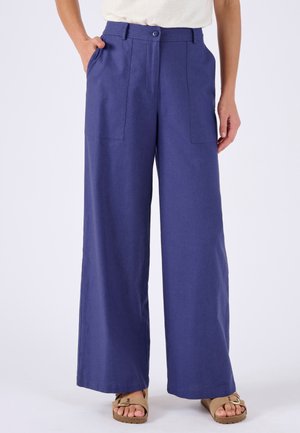 Weit geschnittene blaue Hosen mit aufgesetzten Fronttaschen, getragen mit beigen Sandalen mit Schnalle und einem in die Hose gesteckten weißen Top.