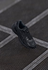 Baskets de sport noires au design épuré, avec une surface texturée, des lacets fins et des accents réfléchissants sur un revêtement à motifs.