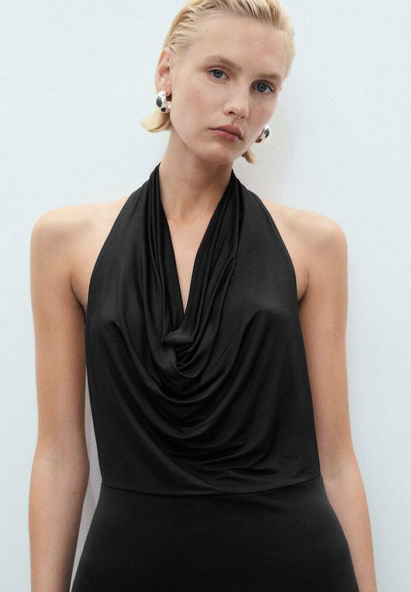 Top negro de halter hecho de una tela suave, con un escote drapeado y una silueta ajustada. Los accesorios incluyen grandes pendientes de plata.