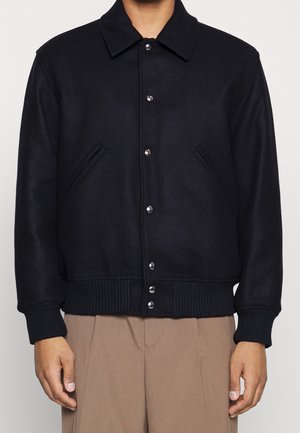 Personne portant une veste noire boutonnée avec des poignets et un bas côtelés, associée à un pantalon marron clair, debout devant un fond uni.