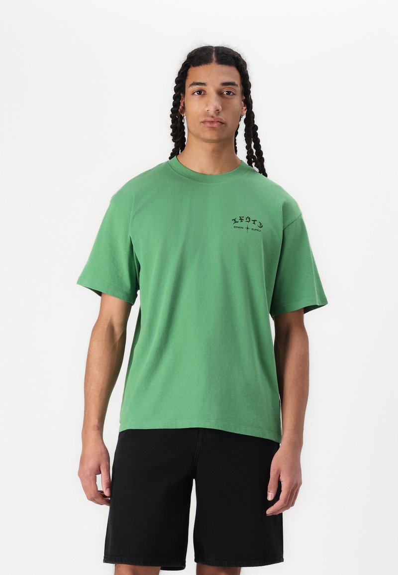 T-shirt verde din bumbac, cu mâneci scurte, având un mic logo negru imprimat pe pieptul stâng. Purtați cu pantaloni scurți din denim negri.