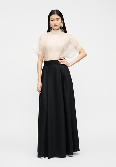 Femme aux longs cheveux noirs portant un haut beige transparent, une jupe longue noire taille haute, et des boucles d'oreilles pendantes, debout avec la main sur la hanche.