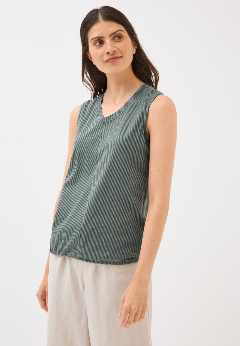 Une femme aux longs cheveux bruns et aux taches de rousseur porte un haut sans manches vert avec un motif à rayures subtiles et un pantalon de couleur claire. Elle regarde sur le côté.